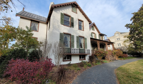 Historic two-story house with green shutters, a porch, and surrounding gardens on a clear day.