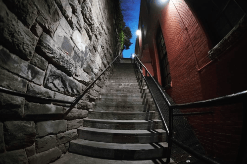A narrow staircase flanked by stone walls, leading up to a dimly lit area with a blue sky above.
