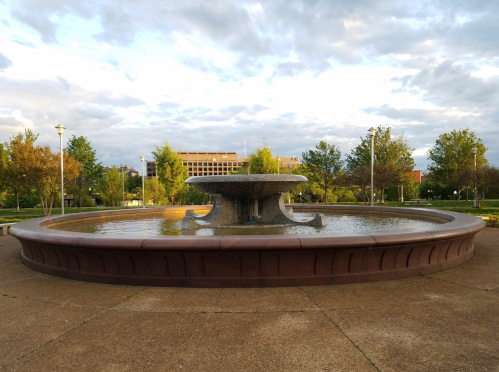 A circular fountain surrounded by trees and grass, with a cloudy sky in the background.