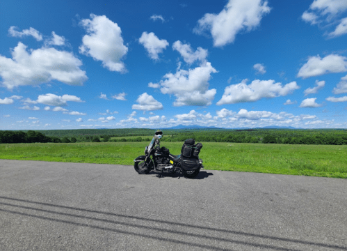 A motorcycle parked on a road with a scenic view of green fields and a blue sky filled with fluffy clouds.