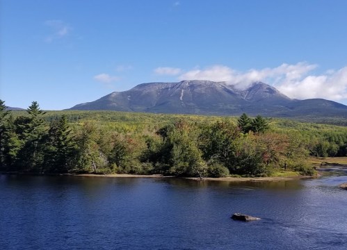 A serene landscape featuring a river, lush trees, and a mountain under a clear blue sky with a few clouds.