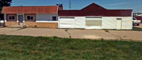 A small, single-story building with a red roof and white walls, featuring a garage and grassy area in front.