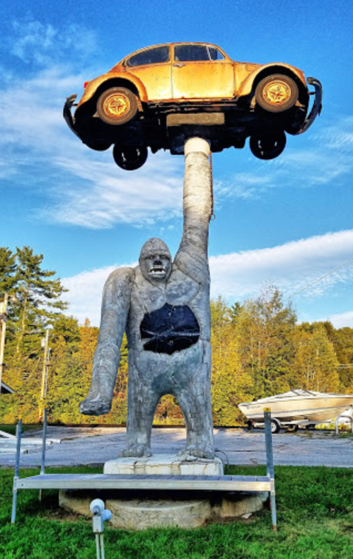 A large statue of a gorilla lifting a vintage car above its head, set against a blue sky and trees in the background.