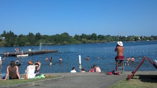 A sunny lakeside scene with people swimming and relaxing on the shore, surrounded by greenery and blue water.