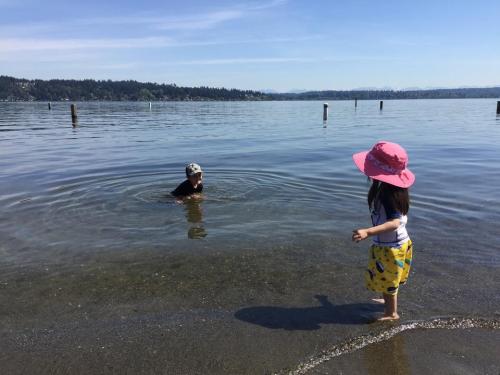 A child in a pink hat stands on the shore while another person swims in a calm lake on a sunny day.