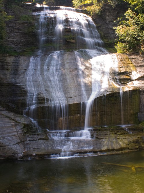 A serene waterfall cascading down rocky cliffs into a calm pool below, surrounded by lush greenery.