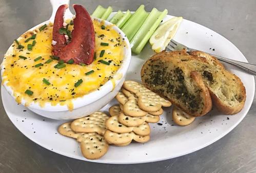 A bowl of lobster mac and cheese topped with lobster, served with celery, lemon, crackers, and garlic bread.