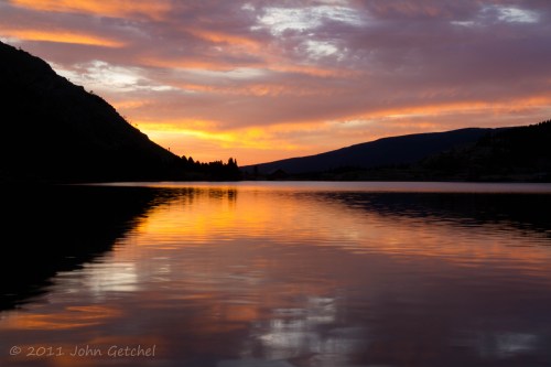 Sunset over a calm lake, with vibrant orange and purple hues reflecting on the water and silhouetted mountains in the background.