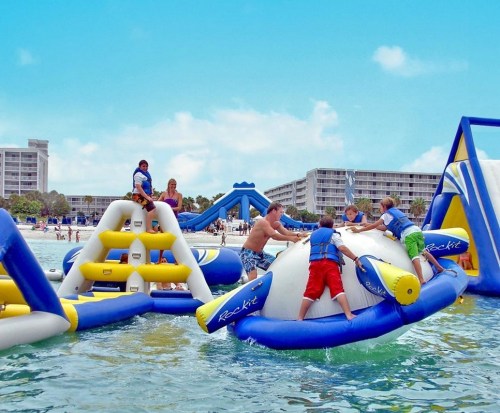 Children and adults play on inflatable water structures at a beach, enjoying a sunny day.