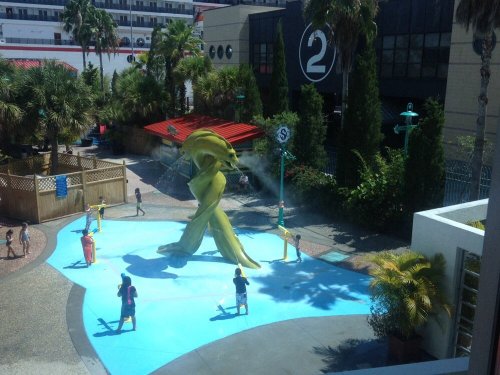 A colorful splash pad with a large yellow sculpture, children playing and spraying water in a sunny outdoor setting.