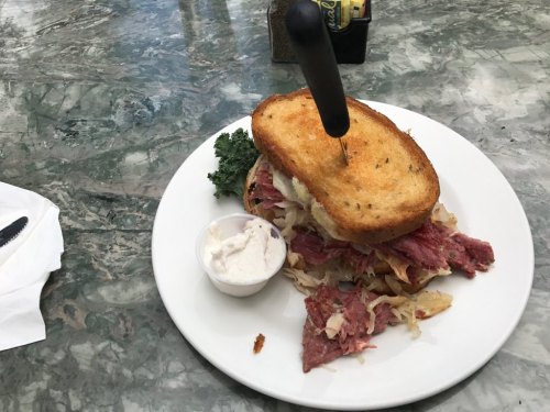 A hearty sandwich with layers of meat and sauerkraut, served with a side of creamy dressing on a marble table.