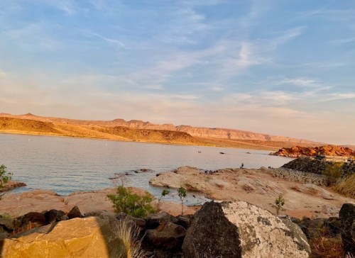 A serene lakeside view with rocky shores, gentle waves, and distant mountains under a clear sky.