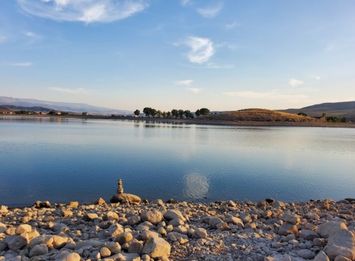 A serene lake scene with calm water, rocky shore, and distant hills under a clear blue sky.