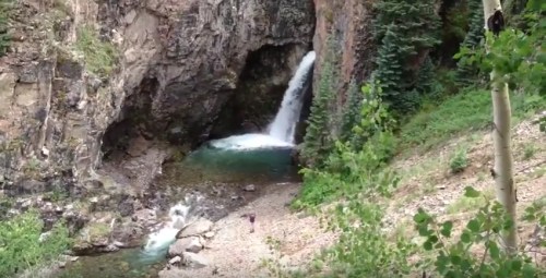 A waterfall cascades into a rocky cave, surrounded by lush greenery and a clear blue pool.