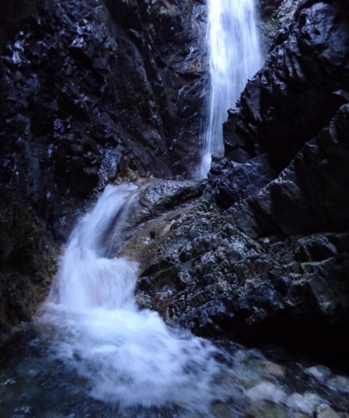 A cascading waterfall flows over rocky terrain, surrounded by dark, rugged cliffs.