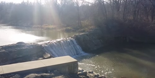 A serene waterfall cascades over rocks into a calm river, surrounded by trees and sunlight.