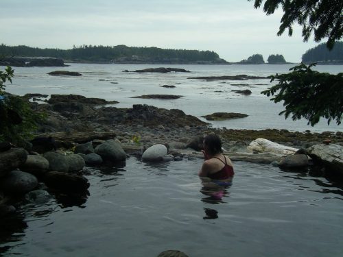 A person relaxes in a natural hot spring by the ocean, surrounded by rocks and trees under a cloudy sky.