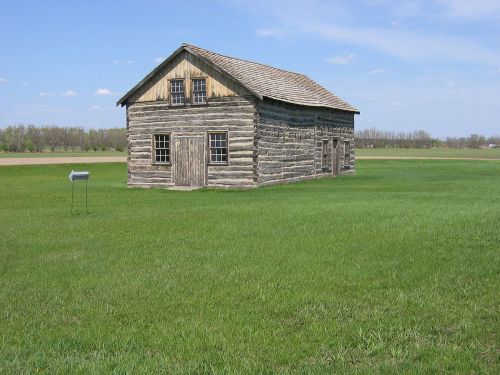 A rustic log cabin stands in a grassy field under a clear blue sky, surrounded by trees in the distance.