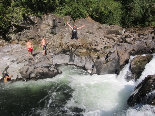A person jumps into a river from a rocky ledge while others watch nearby on a sunny day.