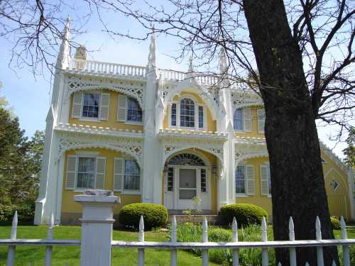 A yellow Gothic-style house with intricate white trim, surrounded by greenery and a white picket fence.