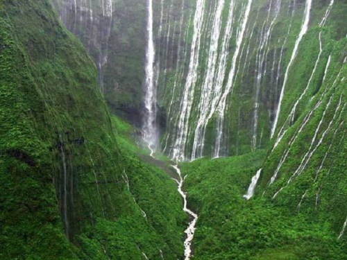 A lush green valley with multiple waterfalls cascading down steep cliffs into a winding river below.