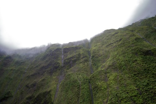 Lush green mountain slopes shrouded in mist, with faint water streams cascading down the rocky surface.