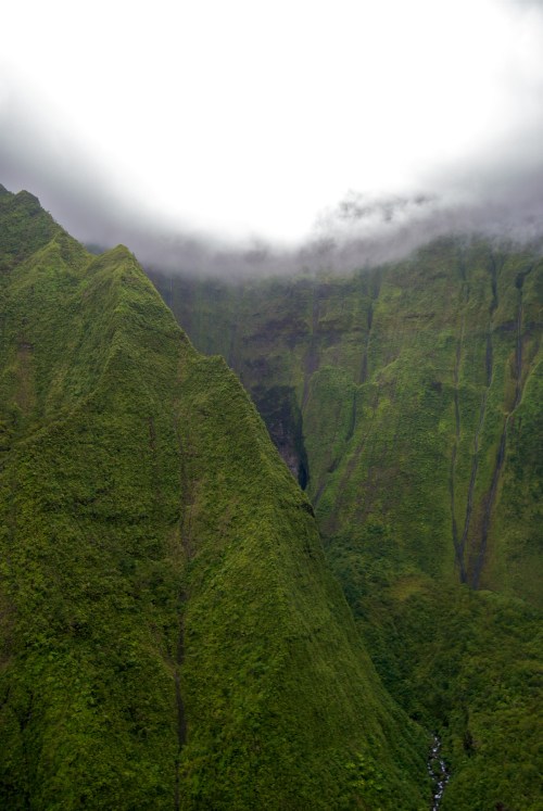 Lush green mountains shrouded in mist, with steep cliffs and a hint of a waterfall in the valley below.
