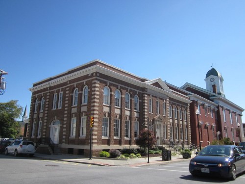 Historic brick building with large windows, next to a clock tower, under a clear blue sky. Cars parked nearby.