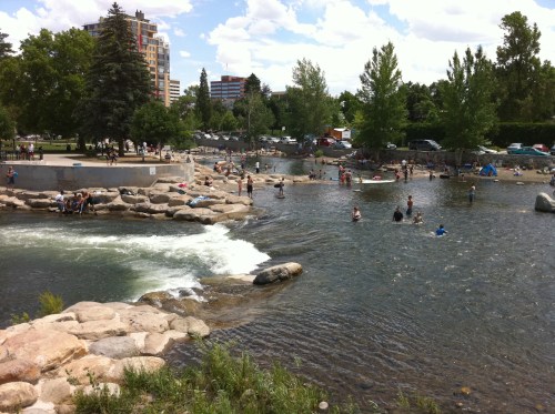 A sunny park scene with people enjoying a river, swimming, and relaxing on rocks, surrounded by trees and buildings.