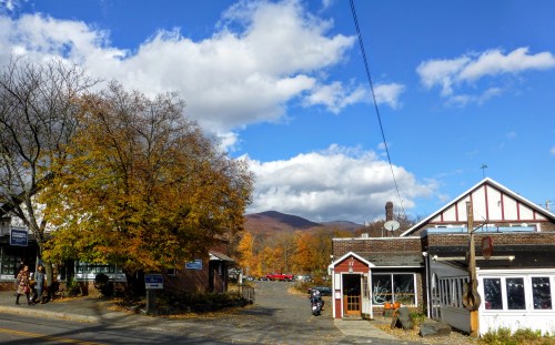 A quaint town scene with autumn trees, blue skies, and mountains in the background, featuring shops and a peaceful atmosphere.