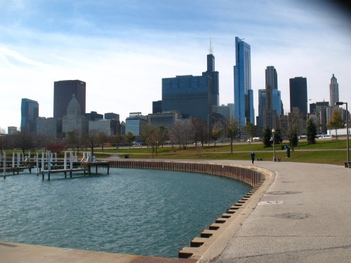 City skyline with modern skyscrapers, a waterfront park, and a calm body of water under a clear blue sky.