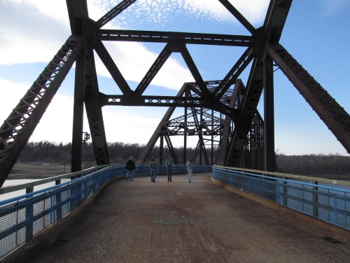 A group of people walking on a metal bridge with a blue railing, surrounded by a scenic landscape and cloudy sky.