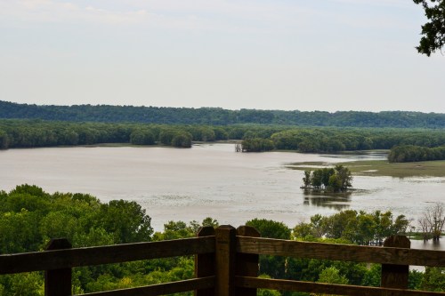 A scenic view of a river surrounded by lush greenery and trees, with a wooden railing in the foreground.