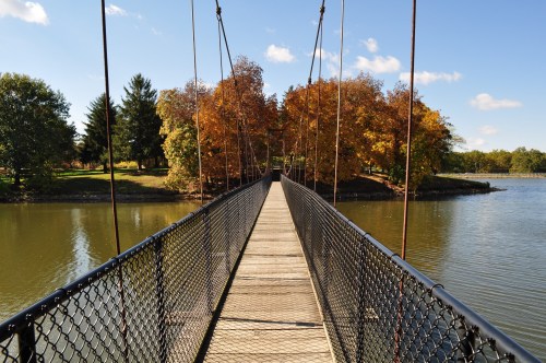 A wooden suspension bridge spans a calm river, surrounded by trees with autumn foliage under a clear blue sky.