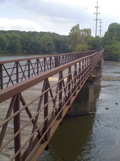 A metal bridge spans a river, surrounded by lush greenery and power lines in the background.