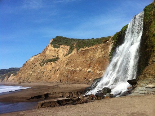 A waterfall cascades down a rocky cliff onto a sandy beach, with blue skies and distant hills in the background.