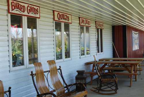 A porch with rocking chairs and picnic tables, featuring signs for baked goods, quilts, and more.