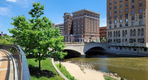 A sunny riverside view featuring a bridge, buildings, and greenery along the water's edge.