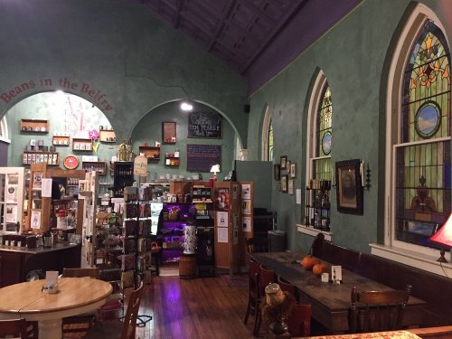Cozy café interior with stained glass windows, wooden tables, and shelves filled with various products. Warm, inviting atmosphere.