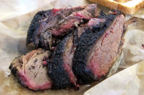 Sliced smoked brisket with a dark crust, served on brown paper with a slice of bread in the background.