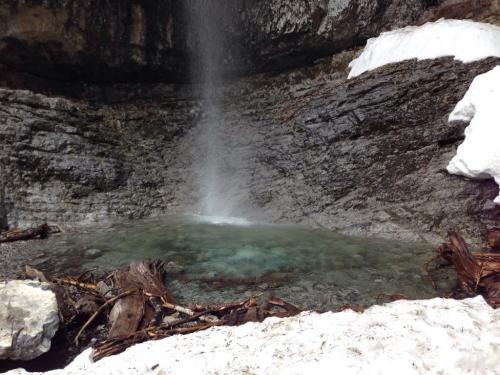 A serene waterfall cascading into a clear pool, surrounded by rocky terrain and patches of snow.