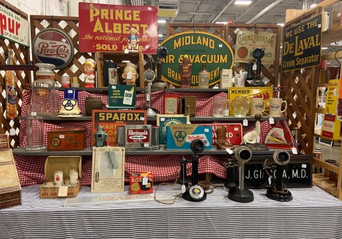 A display of vintage signs, bottles, and antique items on a checkered tablecloth at a market or fair.