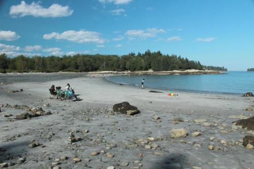 A sandy beach with people relaxing, trees in the background, and a calm blue sea under a clear sky.