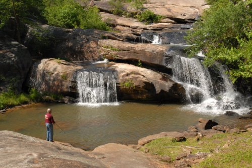 A person stands by a serene waterfall, surrounded by lush greenery and rocky terrain.
