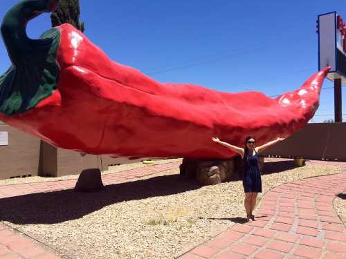 A person stands beside a large red chili pepper sculpture, arms outstretched, under a clear blue sky.