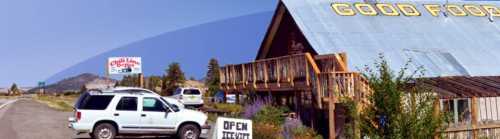 A rustic restaurant with a sign reading "Good Food," surrounded by greenery and parked cars along a highway.