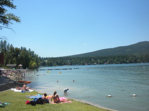 A sunny lakeside scene with people relaxing on the shore, swimming, and enjoying the water against a backdrop of trees and hills.