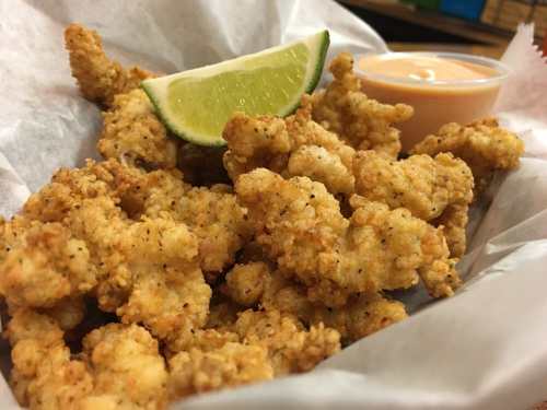 A close-up of crispy fried seafood bites with a lime wedge and a small cup of dipping sauce on a paper-lined tray.