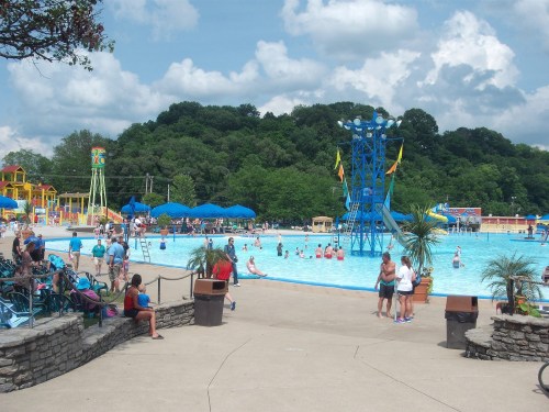 A busy water park with a large pool, slides, and people enjoying the sun under blue skies and fluffy clouds.