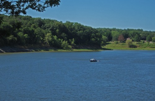 A serene lake scene with a small boat on the water, surrounded by lush green trees and a clear blue sky.